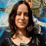 Imagem quadrada de perfil de Thaís Mösken, vestida com uma blusa e um colar, segurando uma carta do jogo Magic. Ao fundo, uma parede azul com pinturas.