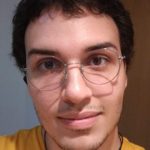Foto quadrada de Luca Nolasco, de óculos, camiseta amarela e um colar. Ao fundo, uma parede branca à esquerda e uma porta bege à direita.